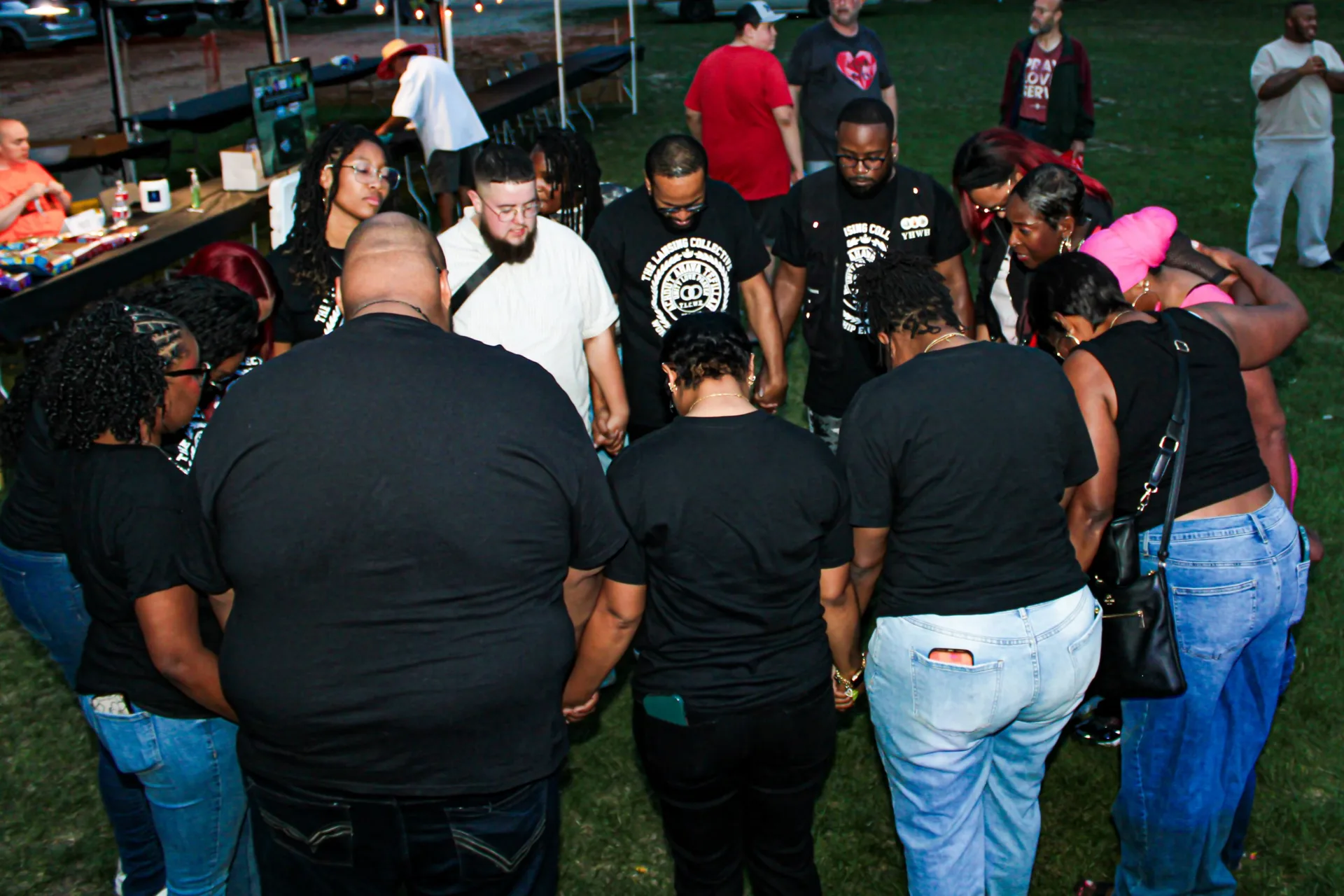 Team members and neighbors holding hands in a circle with heads bowed in prayer on the grass at dusk